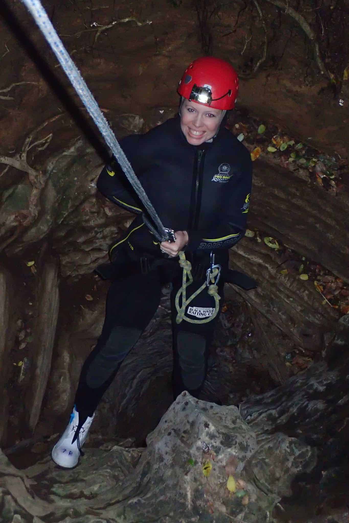 Laura in red helmet and black wetsuit, abseiling into narrow gap, smiling.