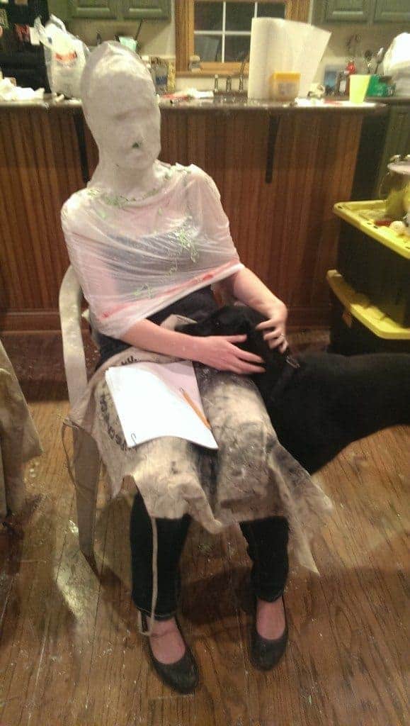 woman seated in chair head wrapped in silicone and plaster, with black Labrador puppy leaning into her lap and soliciting petting